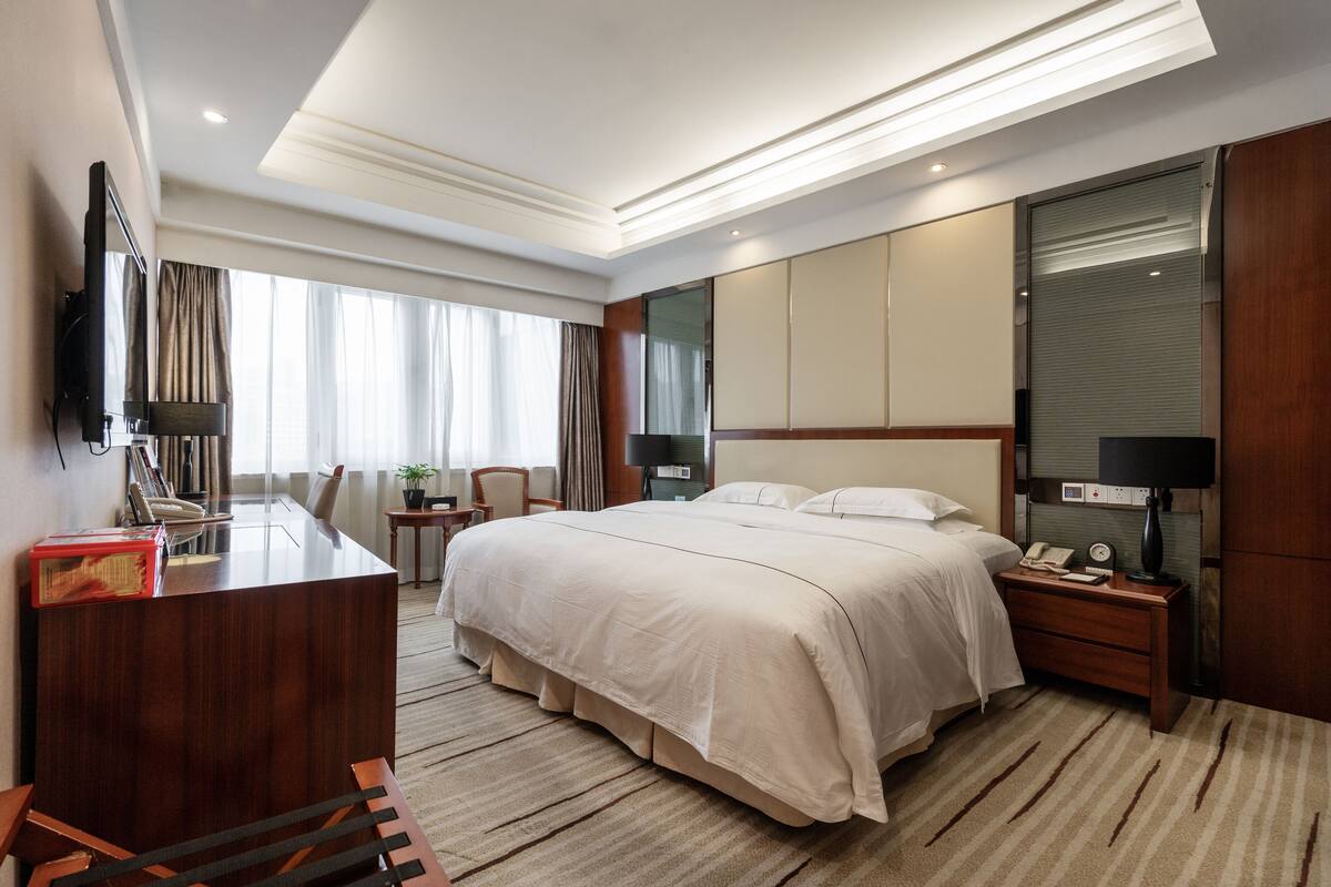Quarto de hotel com uma cama grande, uma escrivaninha com computador, uma TV na parede e uma janela com cortinas.