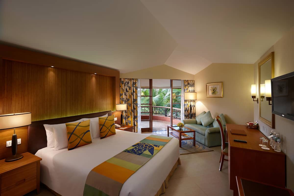 A hotel room with a large bed, a desk, a chair, and a TV. There is a balcony with a view of palm trees.