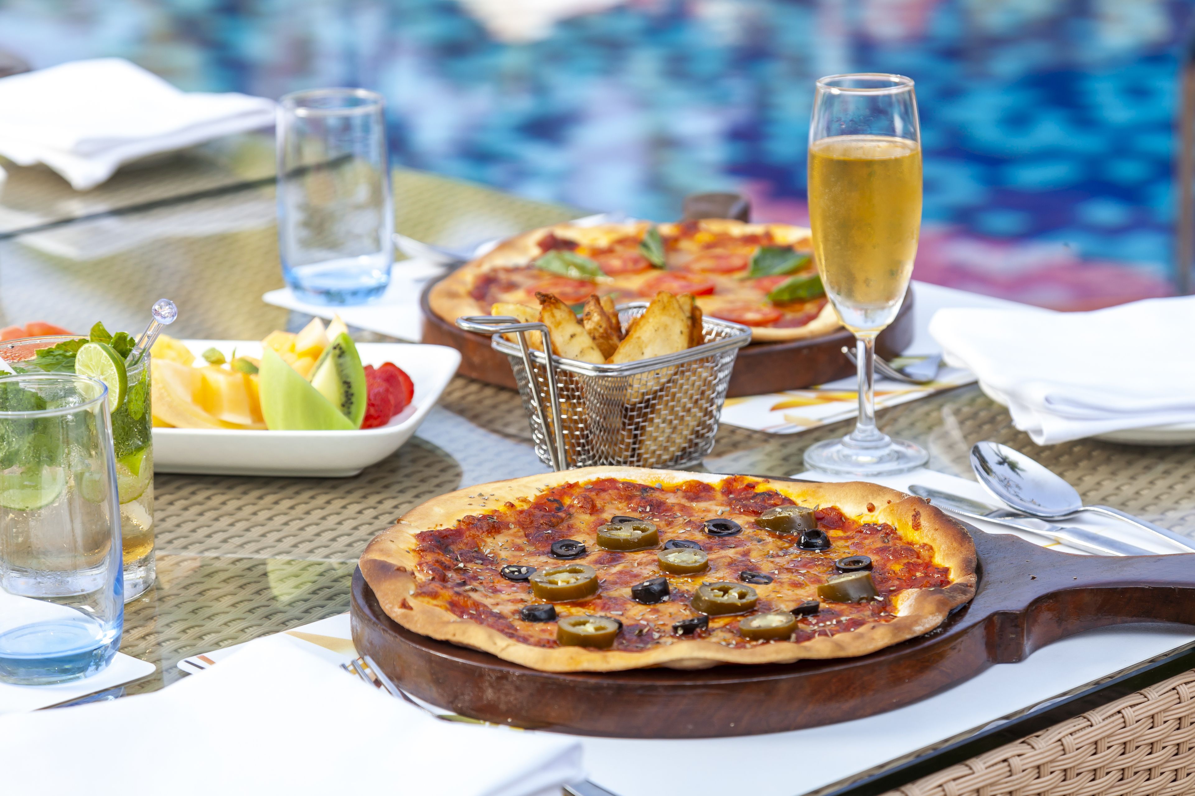 A pizza on a metal tray with olives and jalapeños, a glass of water, and a glass of champagne on a table by a pool.