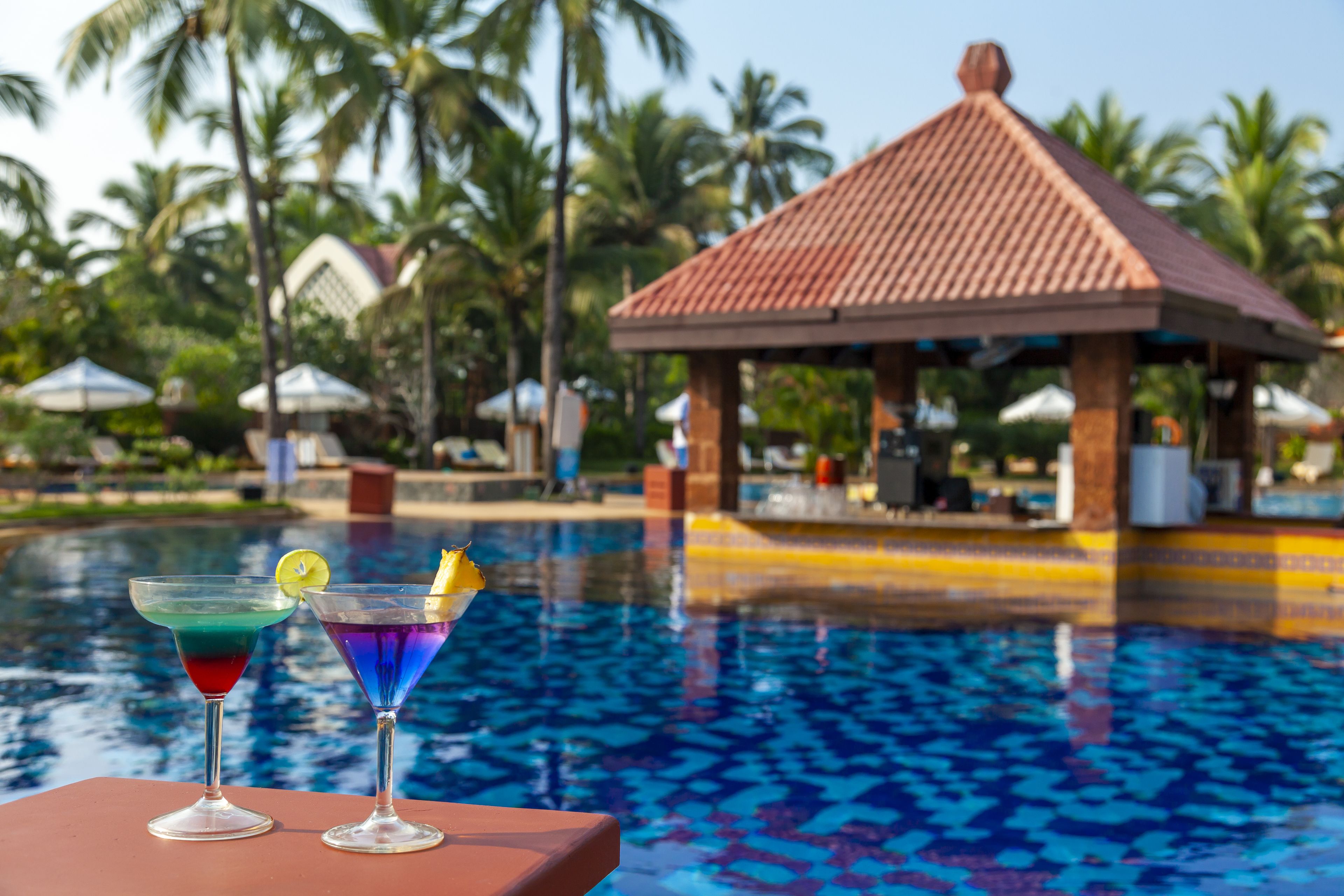 A poolside view with two cocktails on a table, a gazebo in the background, and palm trees.