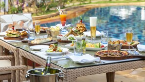 An outdoor dining table with various dishes, drinks, and a bottle of champagne, set against a poolside backdrop. - Caravela Beach Resort (Varca)