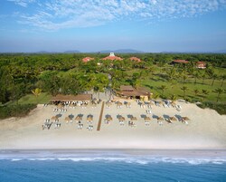 Vista aérea de um resort à beira-mar, com vários edifícios, uma praia arenosa com espreguiçadeiras e vista para o oceano.