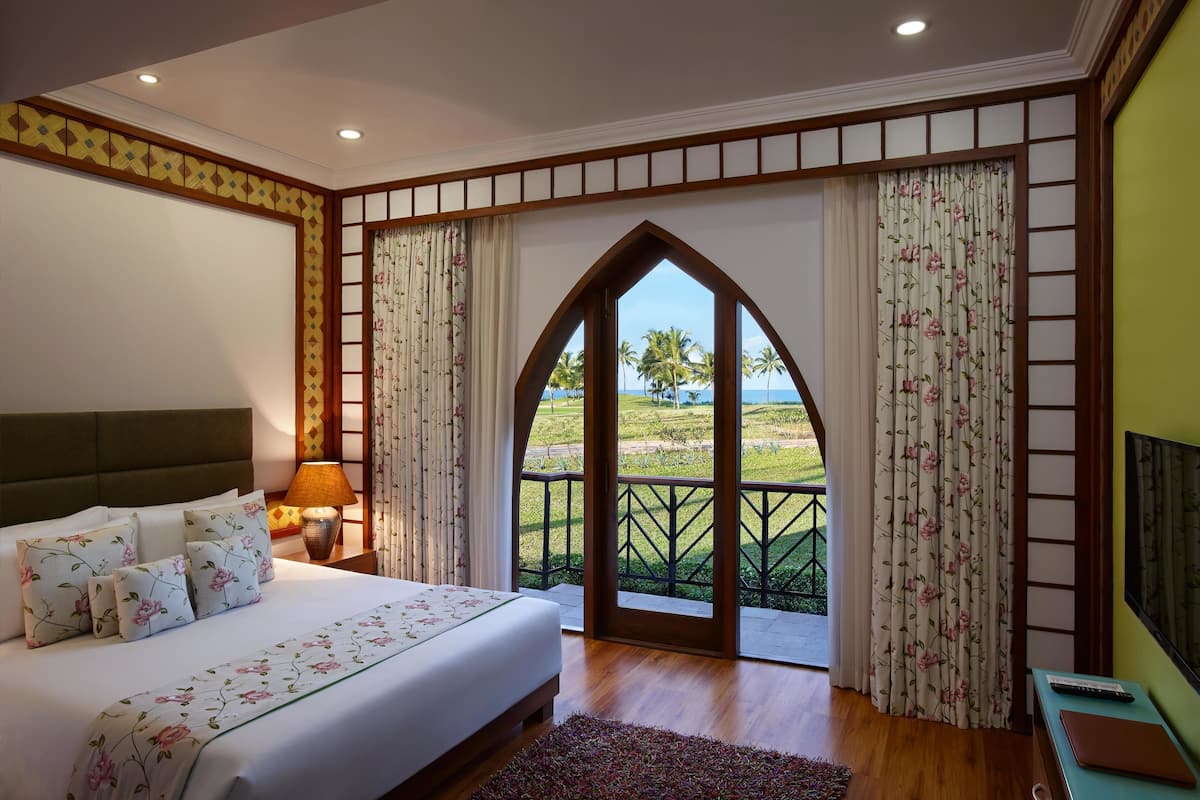A bedroom with a large bed, floral bedding, a nightstand with a lamp, and a view of a balcony with palm trees.