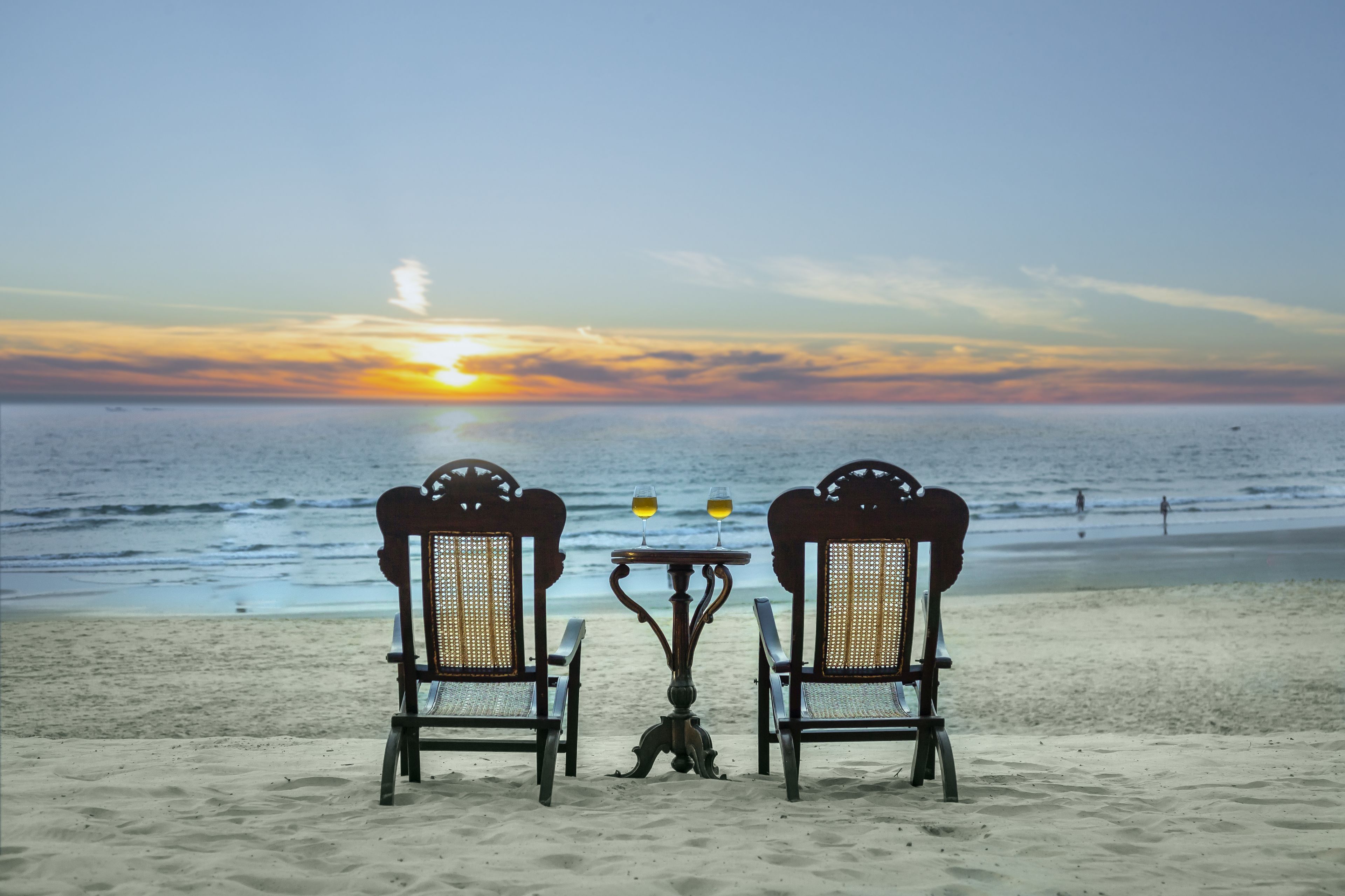 Eine Strandansicht mit zwei Korbstühlen und einem Tisch mit zwei Weingläsern, mit Blick auf den Ozean bei Sonnenuntergang.