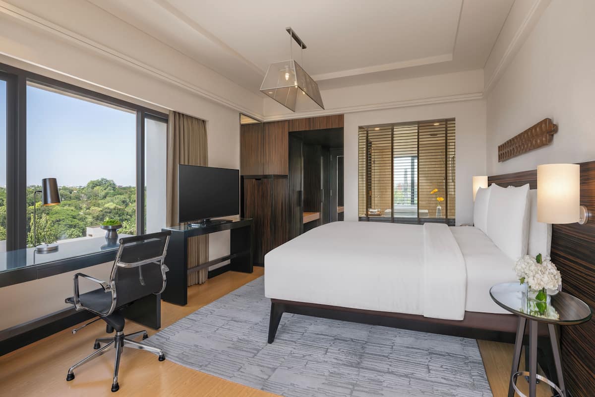 Ein modernes Hotelzimmer mit einem großen Bett, einem Schreibtisch mit Stuhl, einem Fernseher und Blick auf Grün hinter großen Fenstern.