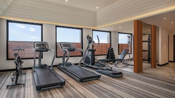 A modern gym with treadmills, elliptical machines, and weight equipment. Large windows provide natural light and views of the sky.