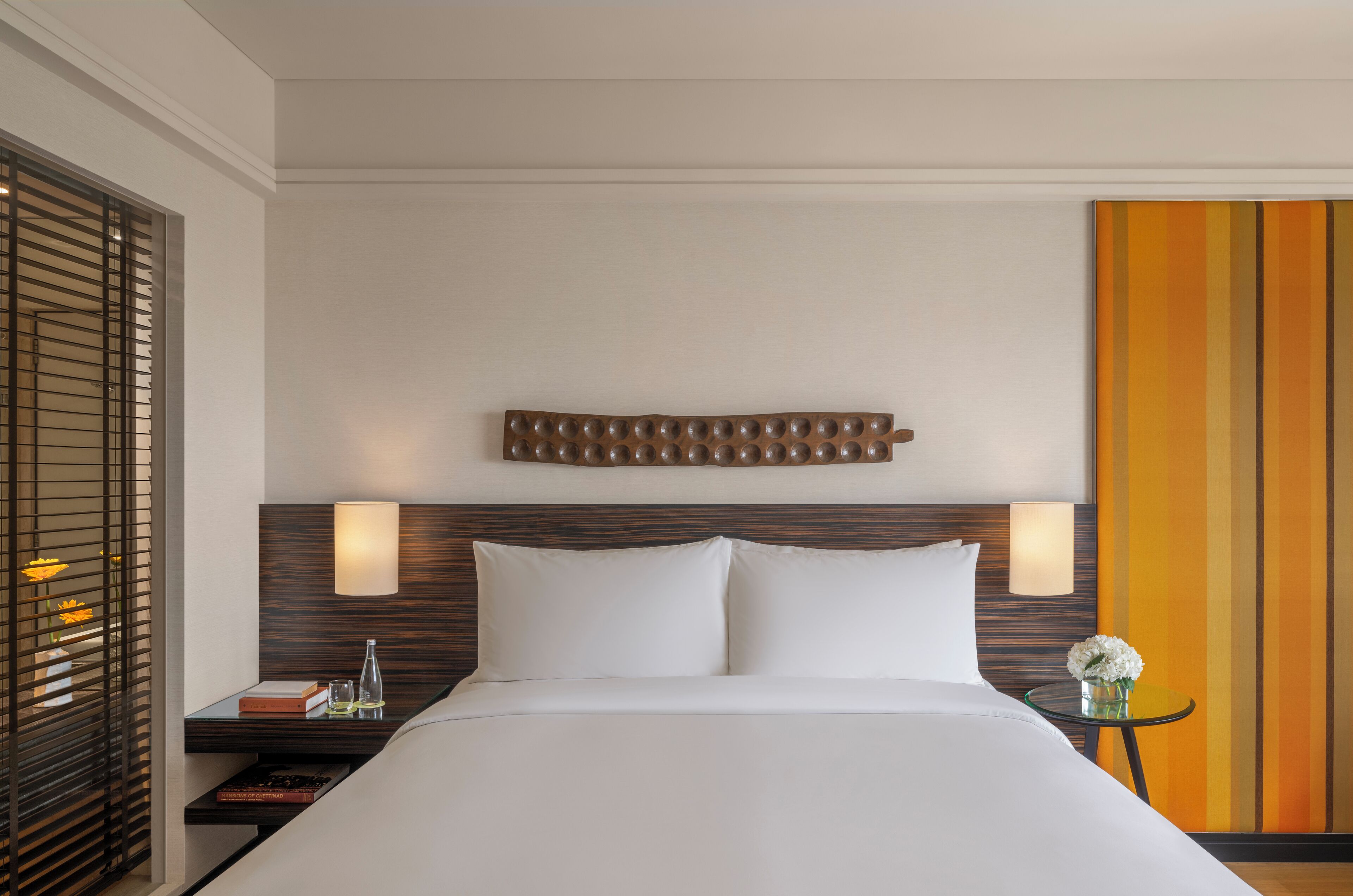 Habitación de hotel con una cama, dos lámparas de noche, una mesita de noche, una pequeña mesa con un jarrón de flores y un panel decorativo en la pared.