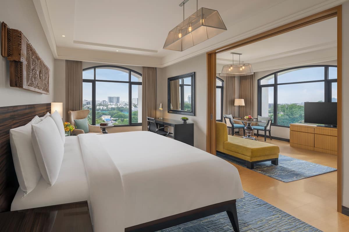 Ein Hotelzimmer mit einem großen Bett, einem Schreibtisch, einem Sessel, einem Fernseher und großen Fenstern mit Blick auf die Stadt.
