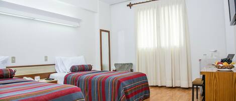 A hotel room with two beds, a desk with a laptop and fruit, and a window with curtains.