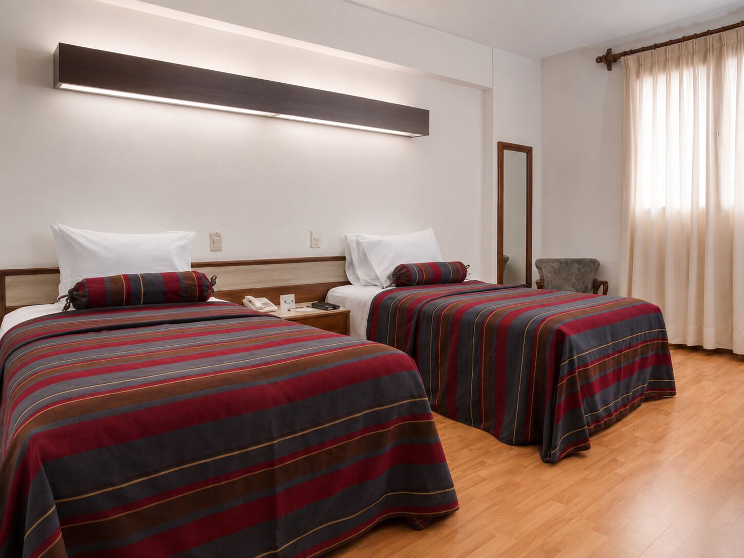 Habitación de hotel con dos camas, una mesita de noche con teléfono, un espejo y una ventana con cortinas.
