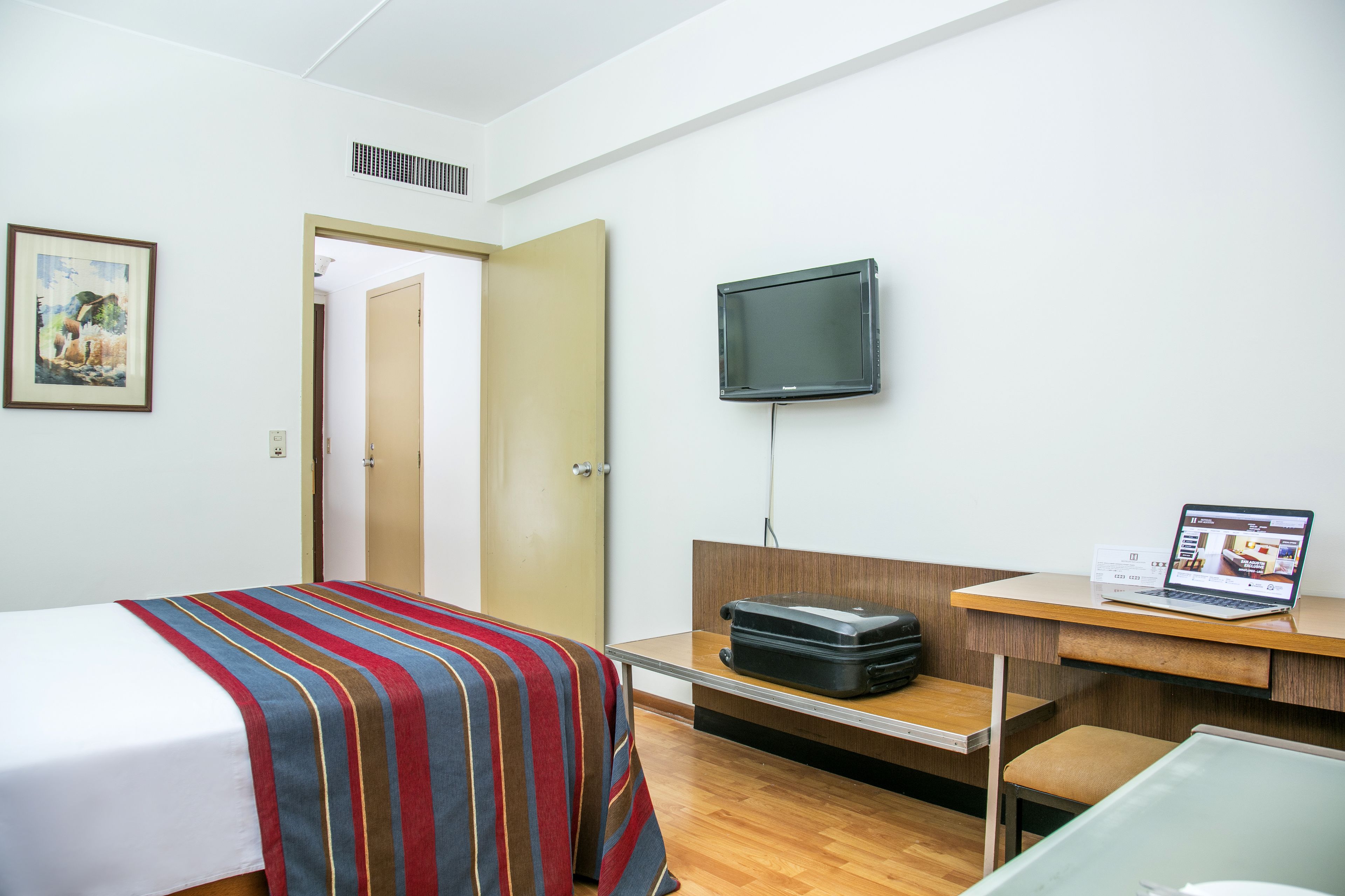 Una habitación de hotel con una cama, un escritorio con una computadora portátil, un televisor y una maleta.