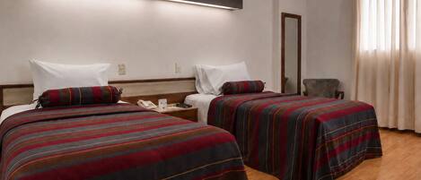 Habitación de hotel con dos camas, una mesita de noche con teléfono, un espejo y una ventana con cortinas.