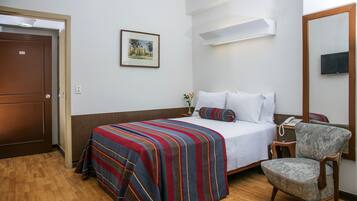 Quarto de hotel com cama, poltrona, mesa de cabeceira com abajur, televisão na parede e porta.