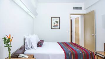 Habitación de hotel con cama, mesita de noche, teléfono, florero con flores, un cuadro en la pared y una puerta que da a otra habitación.