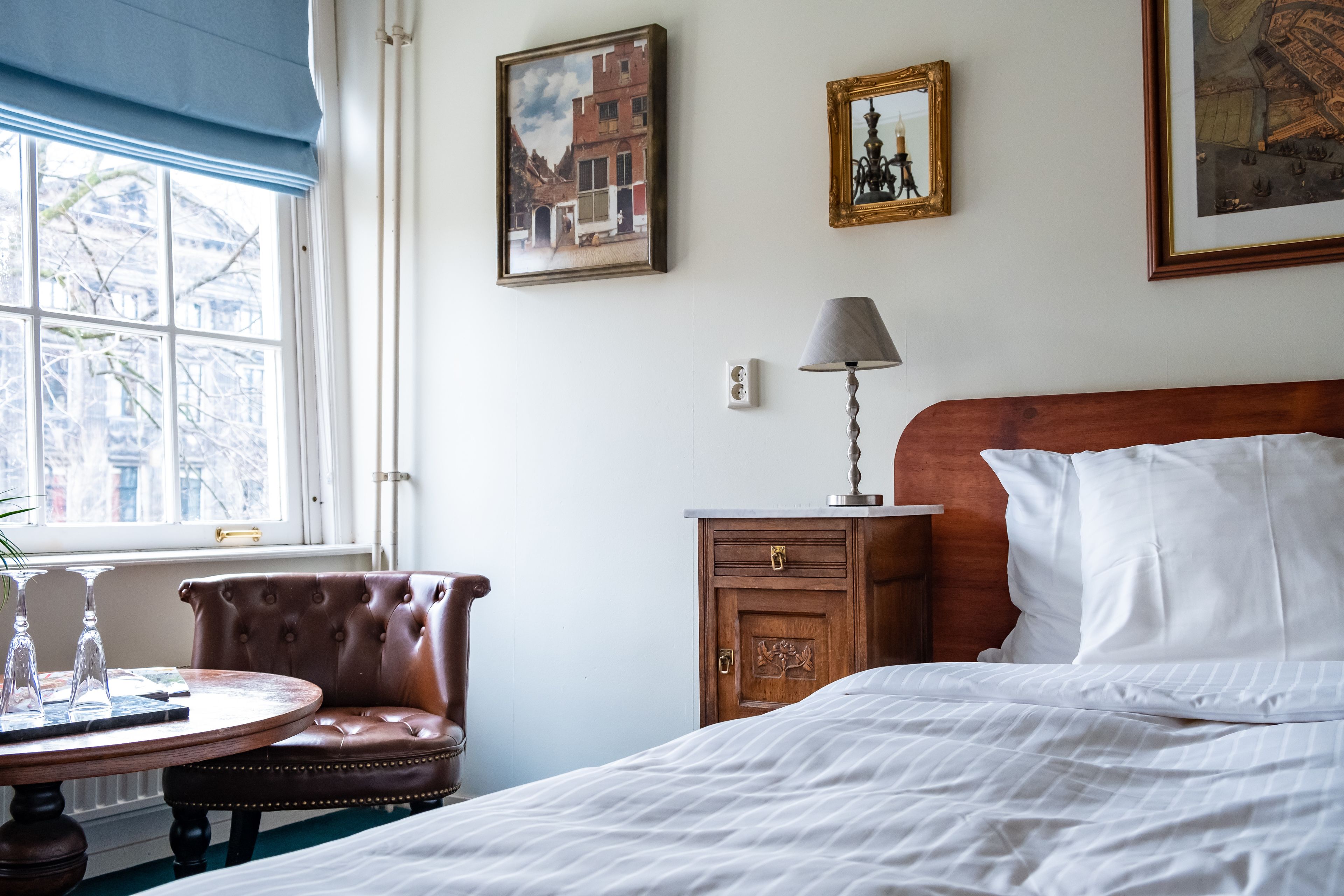 Habitación de hotel con una cama, una mesita de noche de madera, una lámpara, una silla y una mesa redonda con un florero.