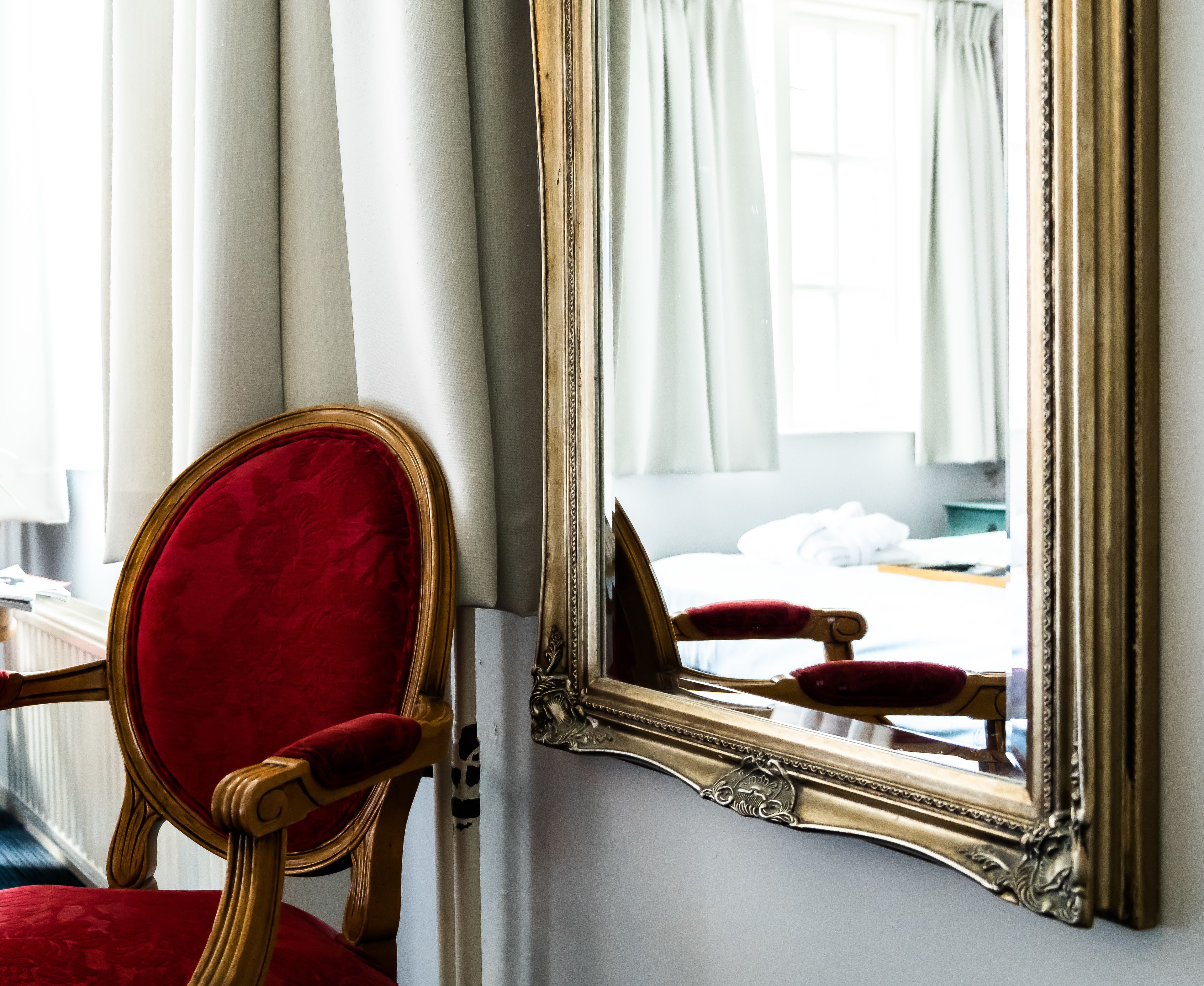 Quarto com um espelho grande, uma poltrona estofada em vermelho e uma cama com roupa de cama branca.