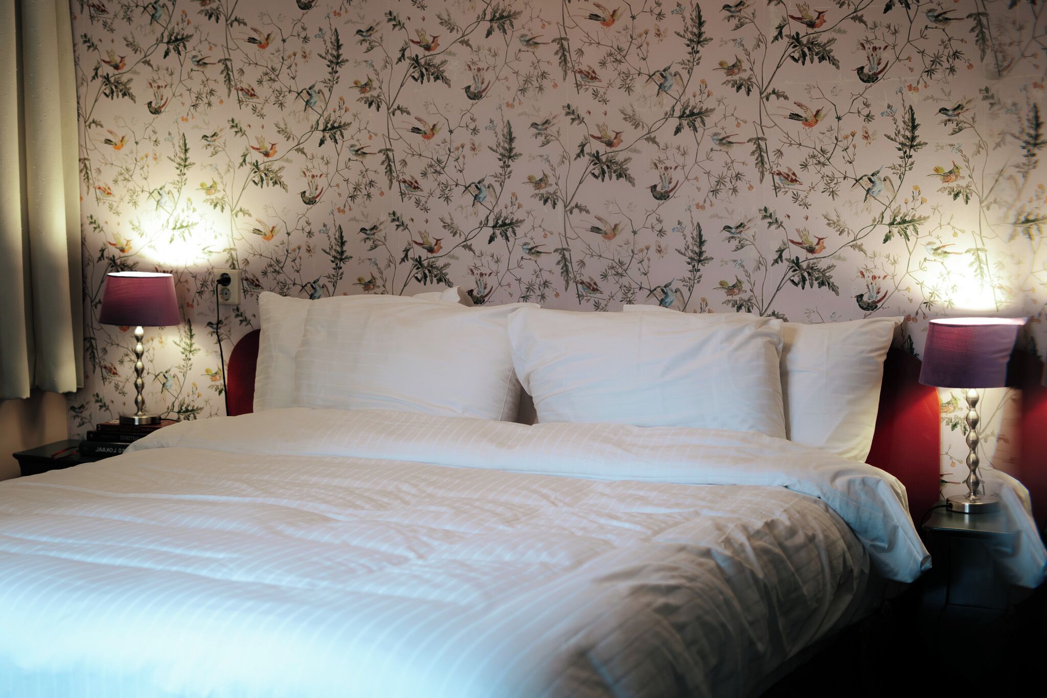A neatly made bed with white linens, two bedside lamps with red shades, and floral wallpaper on the wall.