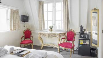 Habitación de hotel con cama, dos sillones tapizados en rojo, una mesita con un jarrón, cafetera y un espejo.