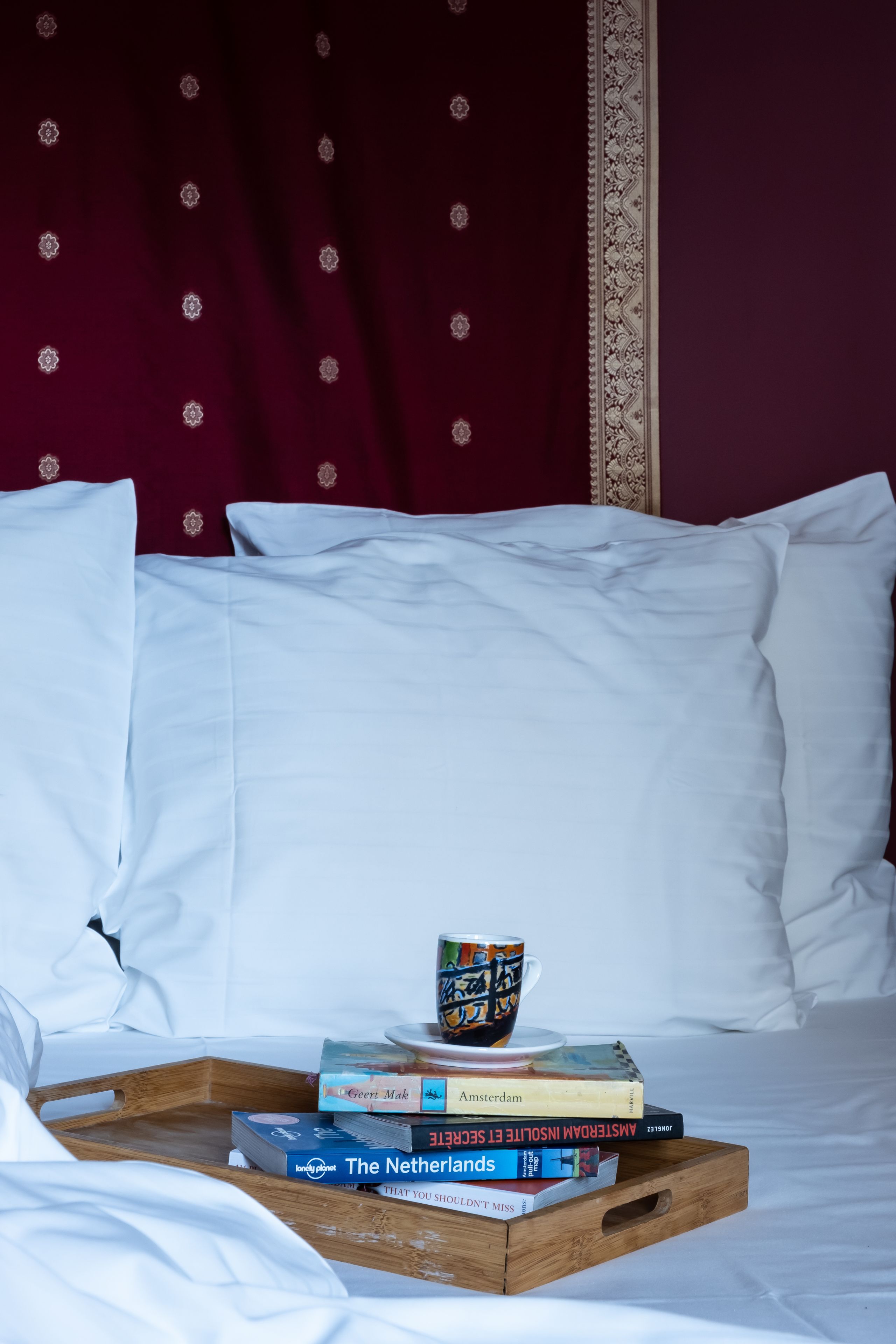 Un lit bien fait, avec du linge de lit blanc, et un plateau en bois posé dessus, sur lequel se trouvent des livres et une tasse.