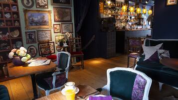 A cozy bar area with wooden furniture, a variety of framed artwork on the walls, and a selection of bottles and glasses on shelves.