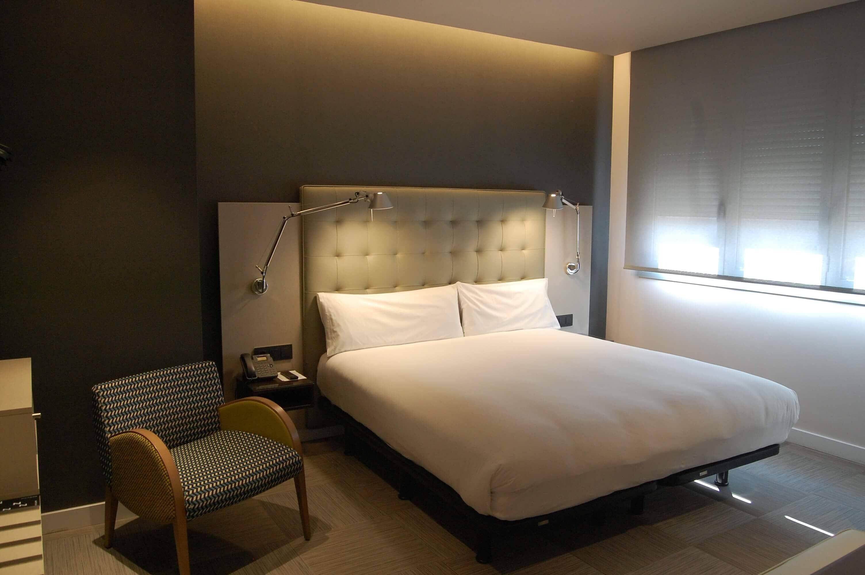 A modern hotel room with a large bed, a bedside lamp, a chair, and a window with blinds.