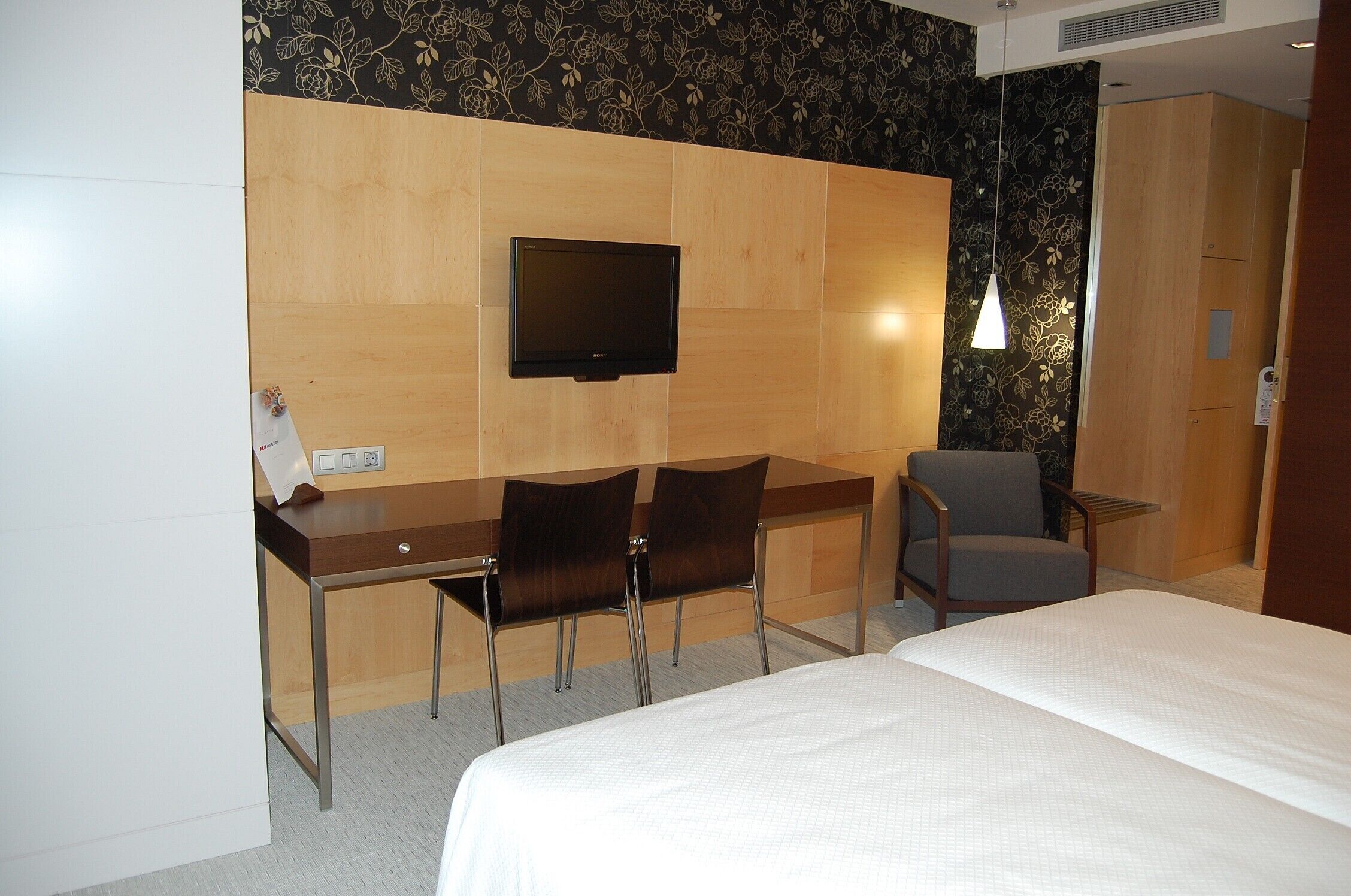 Habitación de hotel con cama, escritorio con dos sillas, televisor y lámpara de pared.