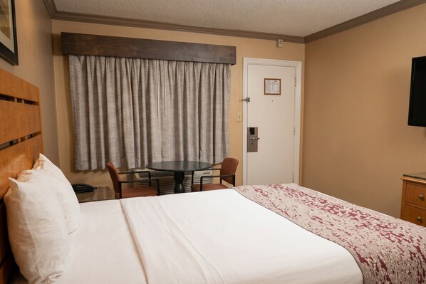 A hotel room with a bed, a small table with chairs, a wooden headboard, a door, and a television on the wall.
