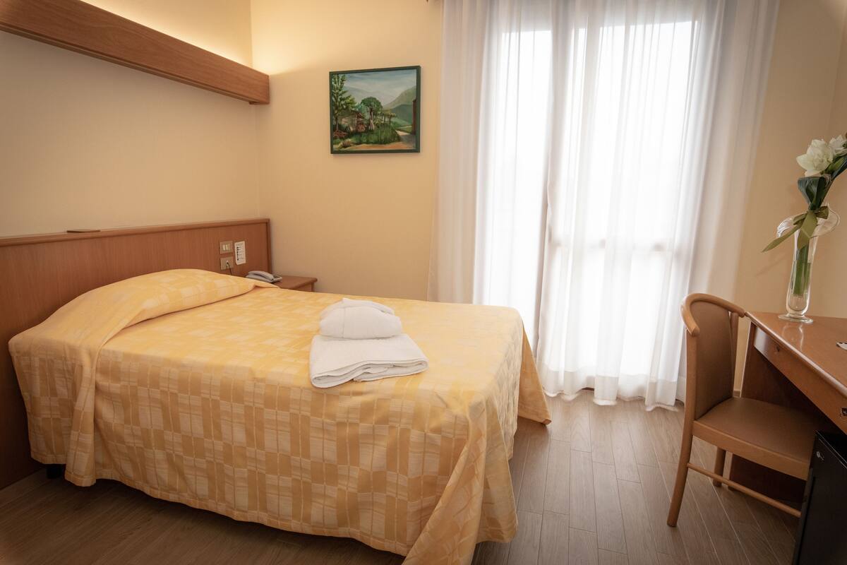 A hotel room with a bed, a desk, a chair, a painting on the wall, and a window with curtains.