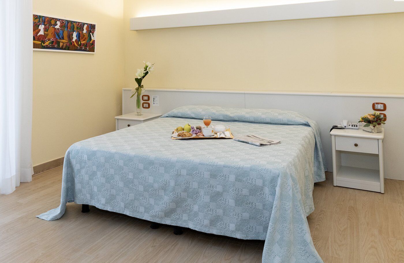 A bedroom with a bed, bedside tables, a nightstand, a painting, and a vase with flowers.