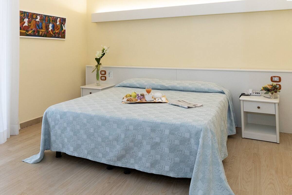 A bedroom with a bed, bedside tables, a nightstand, a painting, and a vase with flowers.