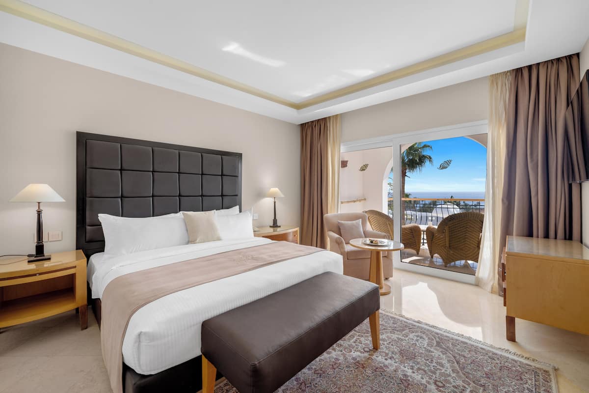 A hotel room with a large bed, a bench, a small table, a chair, and a view of the sea.