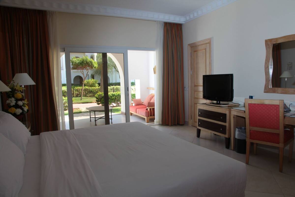 A hotel room with a bed, a desk with a TV, a chair, and a view of a garden through a glass door.