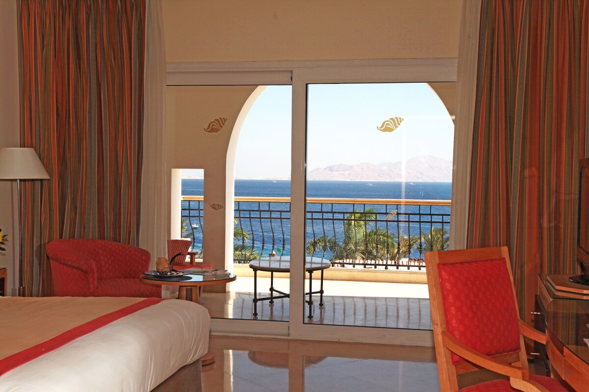 A hotel room with a bed, a red chair, a small table, and a view of the sea through a glass door.