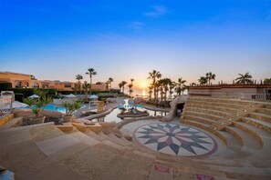 A resort pool area with steps leading to a seating area and a sunken pool, surrounded by palm trees and a sunset view. - Savoy Sharm El Sheikh (Sharm El Sheikh)