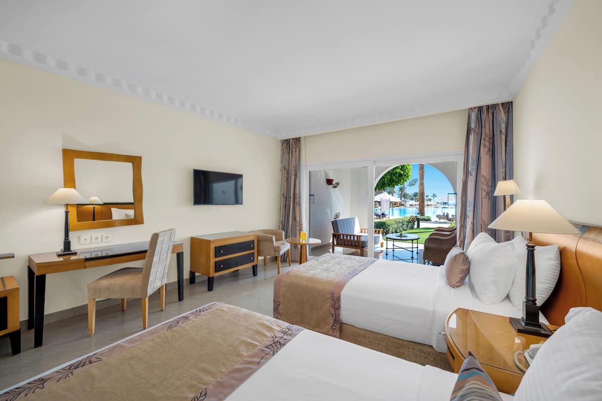 A hotel room with two beds, a desk, a chair, a TV, and a view of the pool.