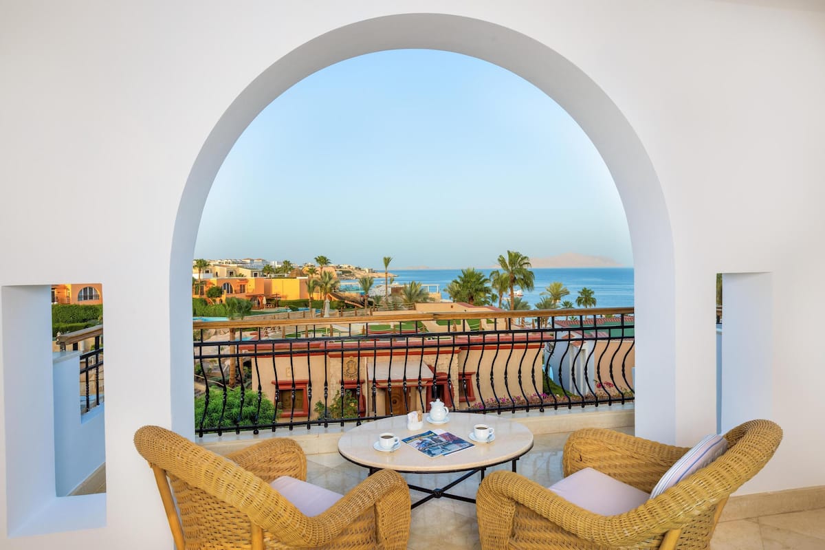 A balcony with wicker chairs, a small table, and a view of the sea and buildings.