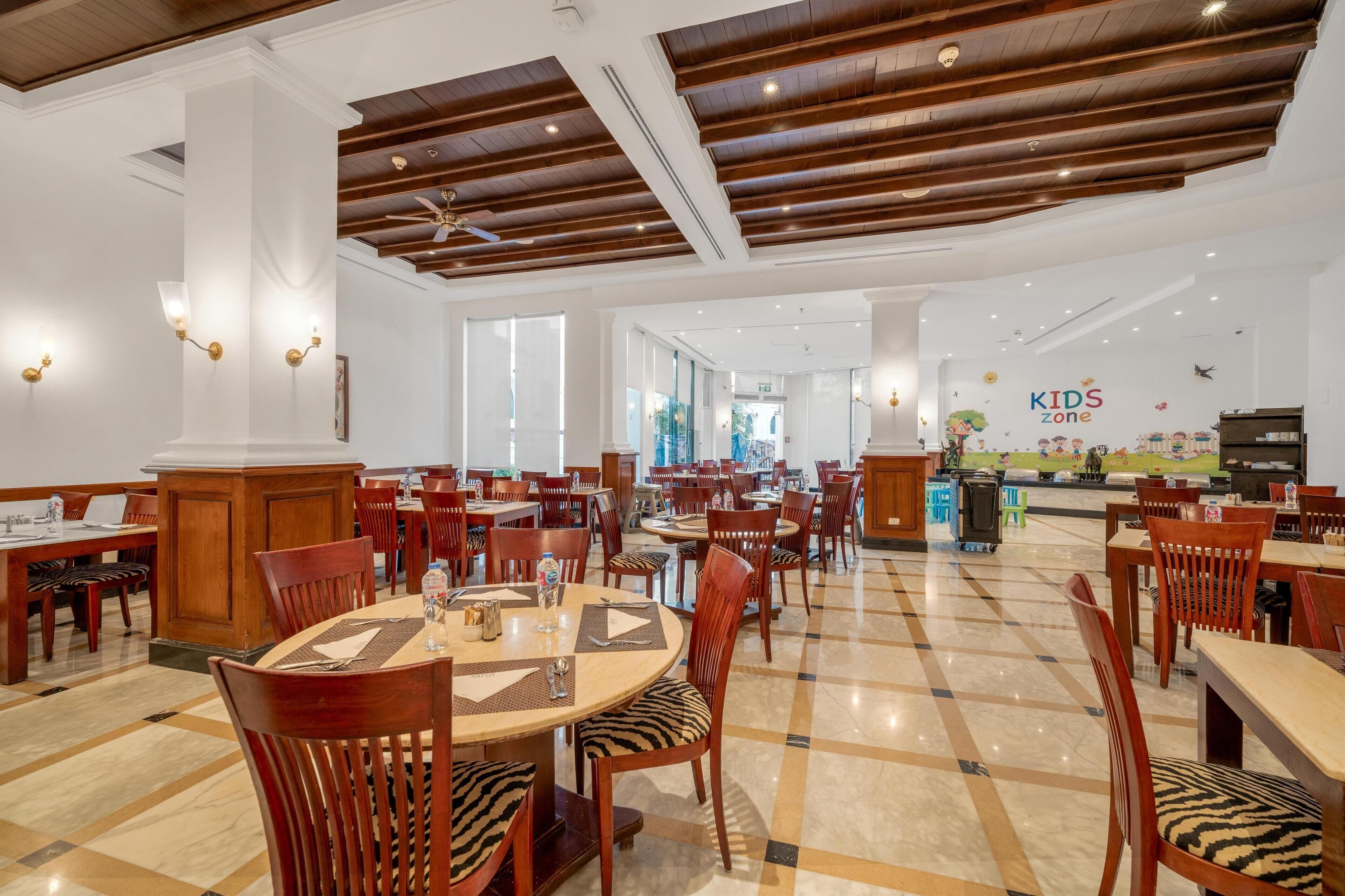 A spacious dining area with wooden tables and chairs, a children's play zone in the background, and a modern ceiling design.