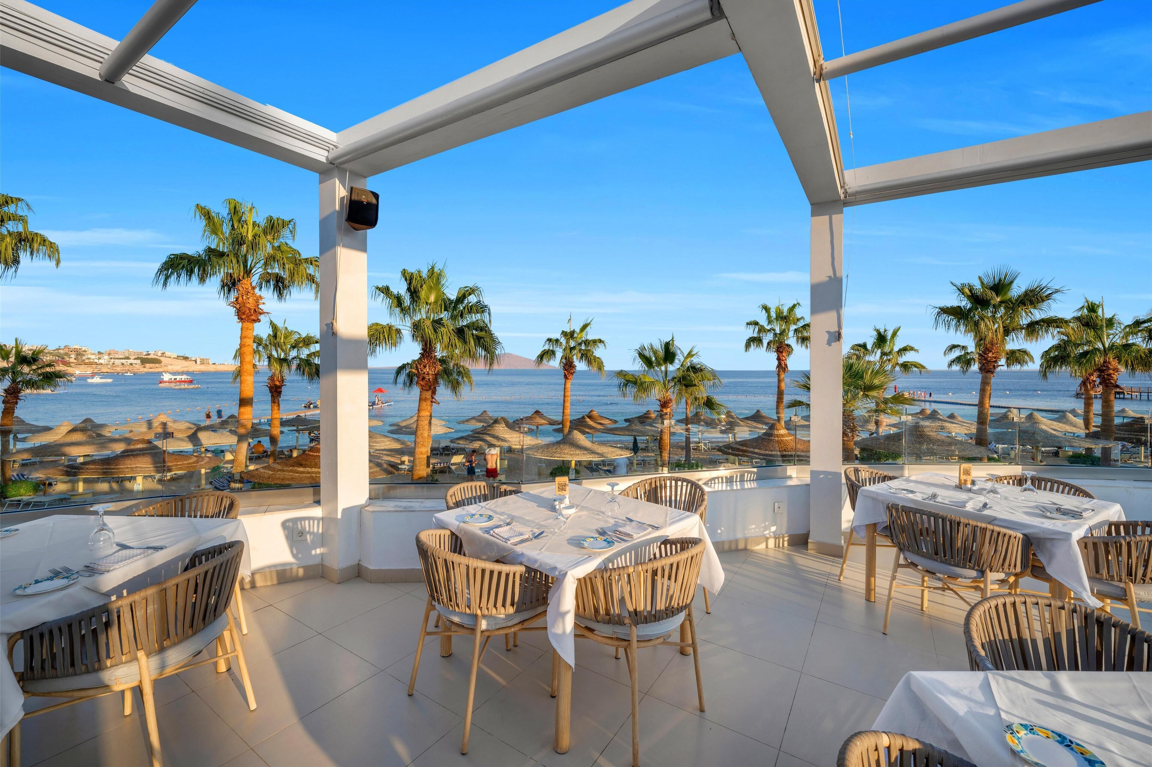 Un restaurante con terraza al aire libre con vistas a una playa, con palmeras y sombrillas.