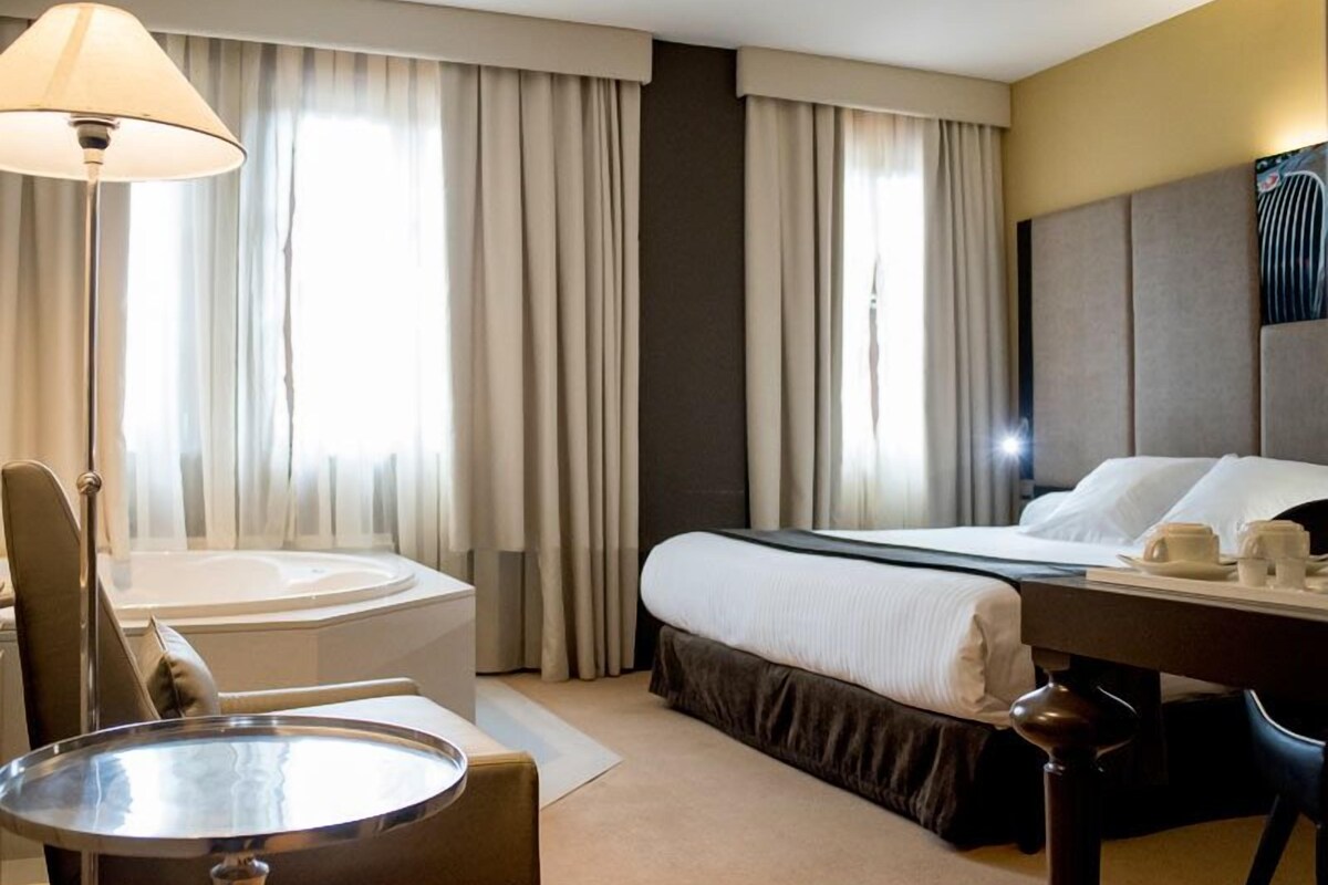 A hotel room with a bed, a desk, a chair, a lamp, and a large window with curtains.