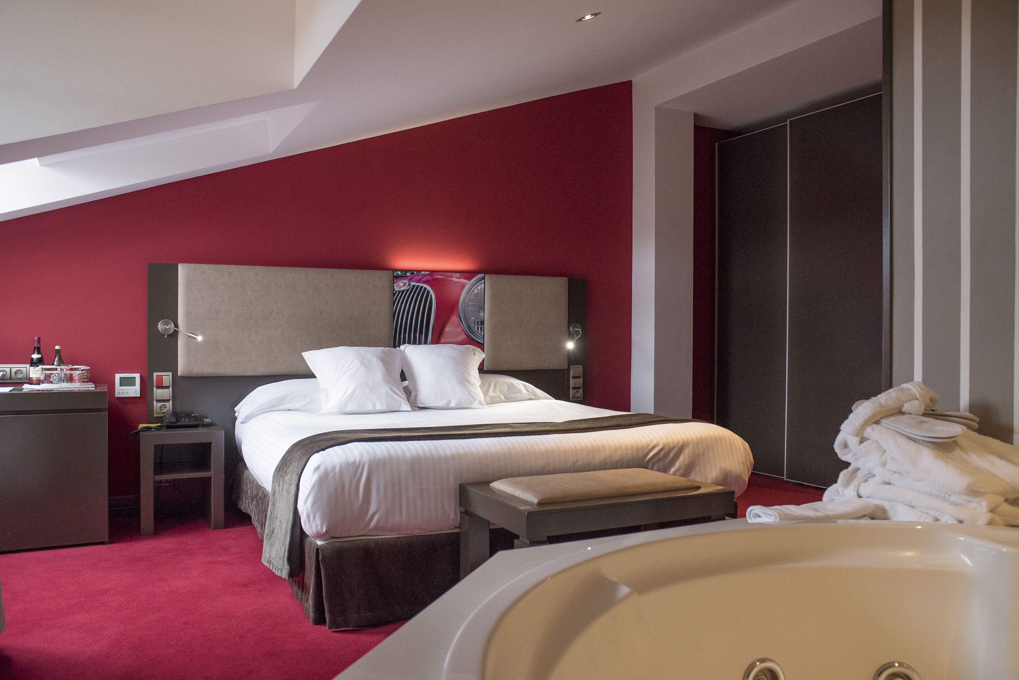 A modern hotel room with a large bed, a bedside table, a desk, a red accent wall, a large mirror, and a bathtub.