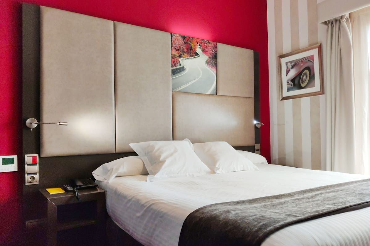 A hotel room with a large bed, a nightstand with a phone and a notepad, a wall clock, and a painting of a winding road.