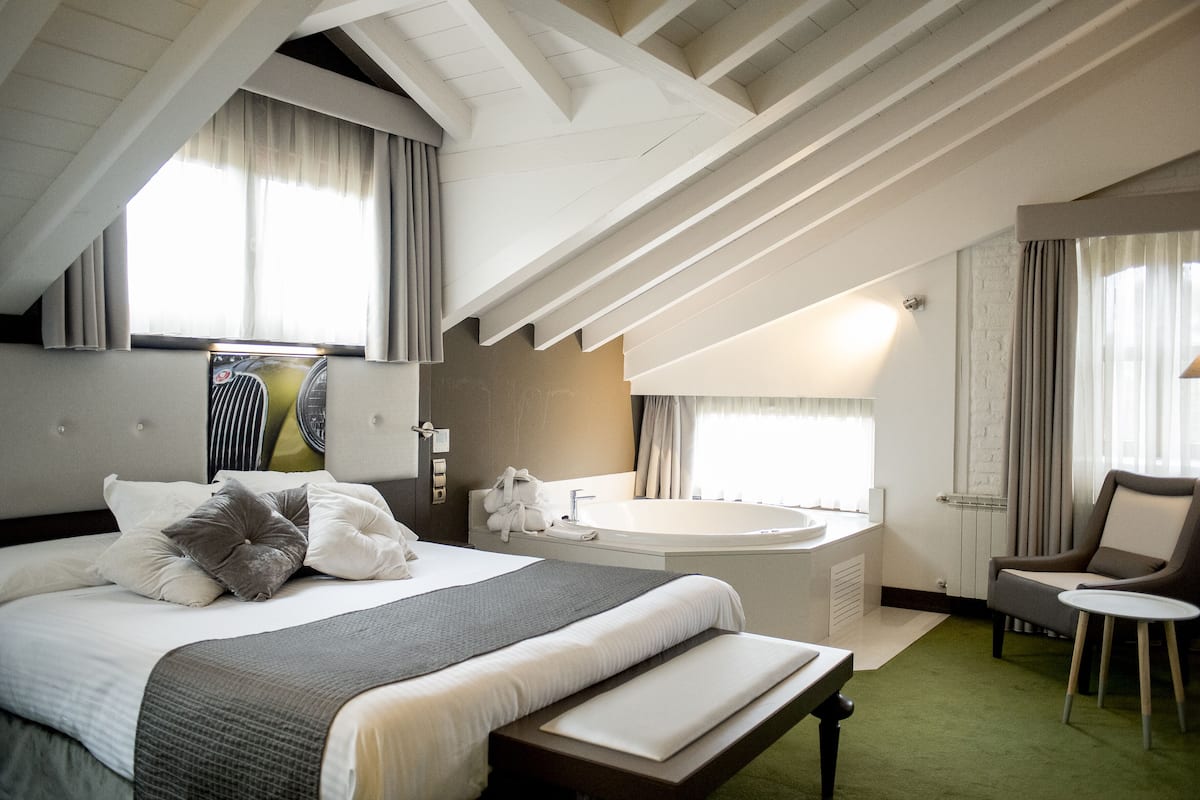 A hotel room with a large bed, a sitting area with a chair and a small table, a bathtub, and a large window with curtains.