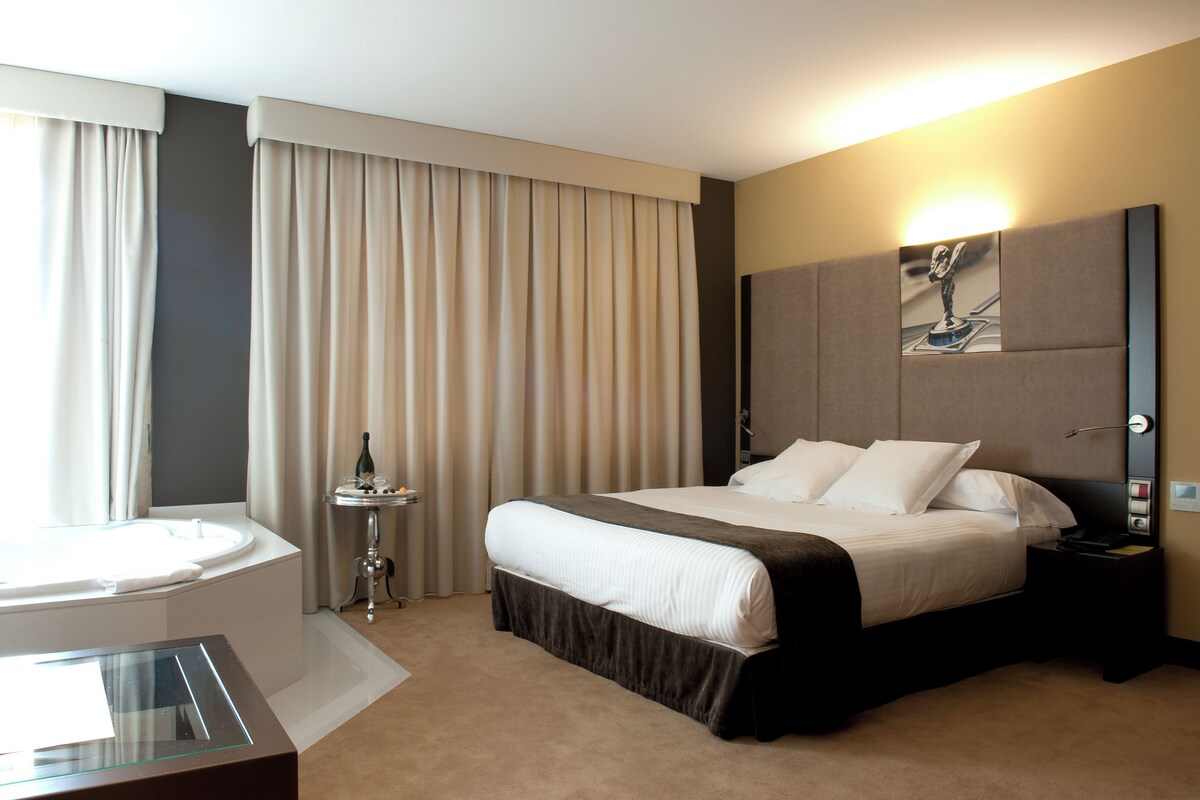 Una habitación de hotel con una cama grande, una mesita de noche, una lámpara de noche, una obra de arte en la pared y una ventana con cortinas.