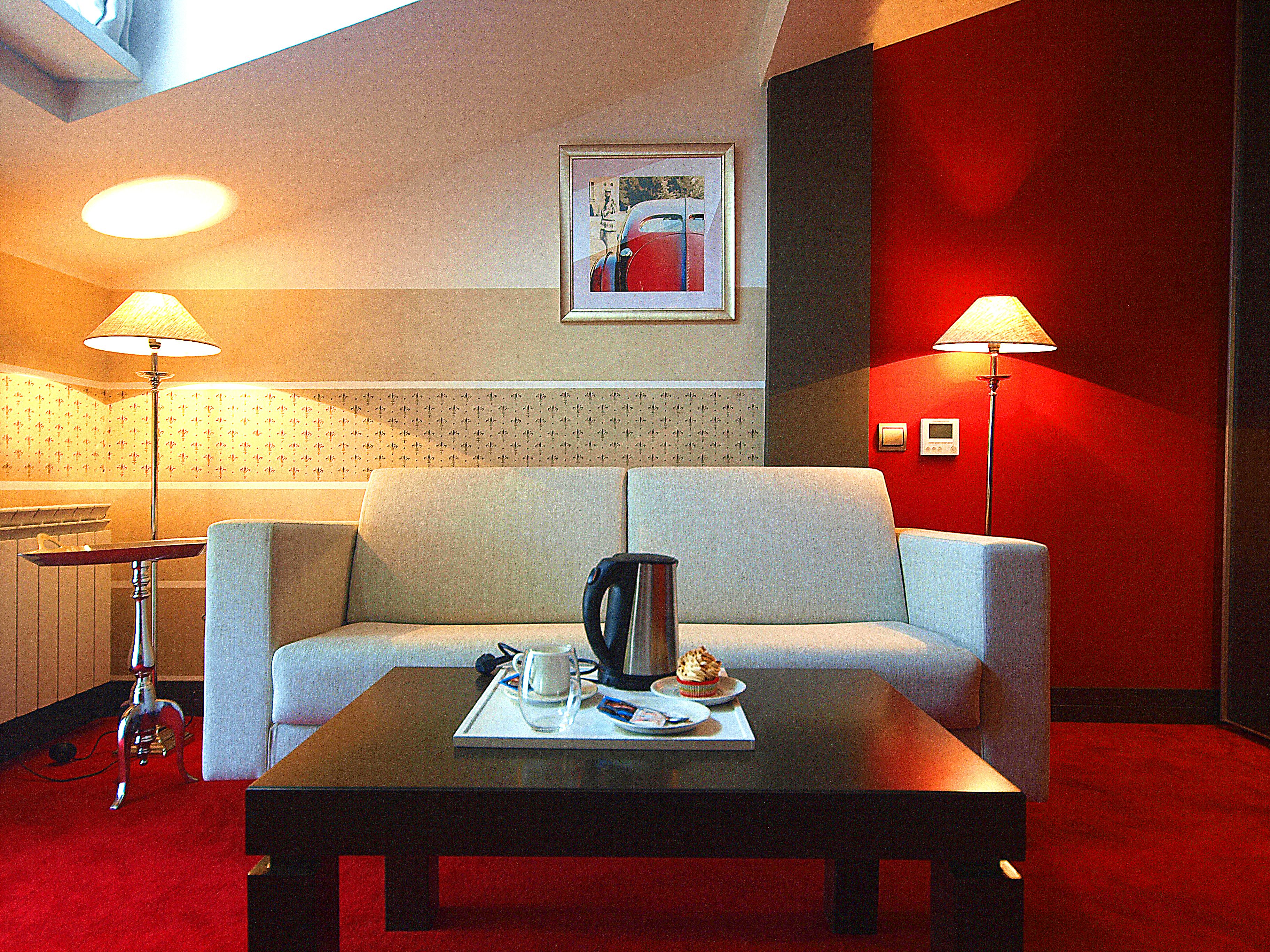 Habitación de hotel con sofá, una mesa de centro con una taza y un pastel, dos lámparas y un cuadro en la pared.