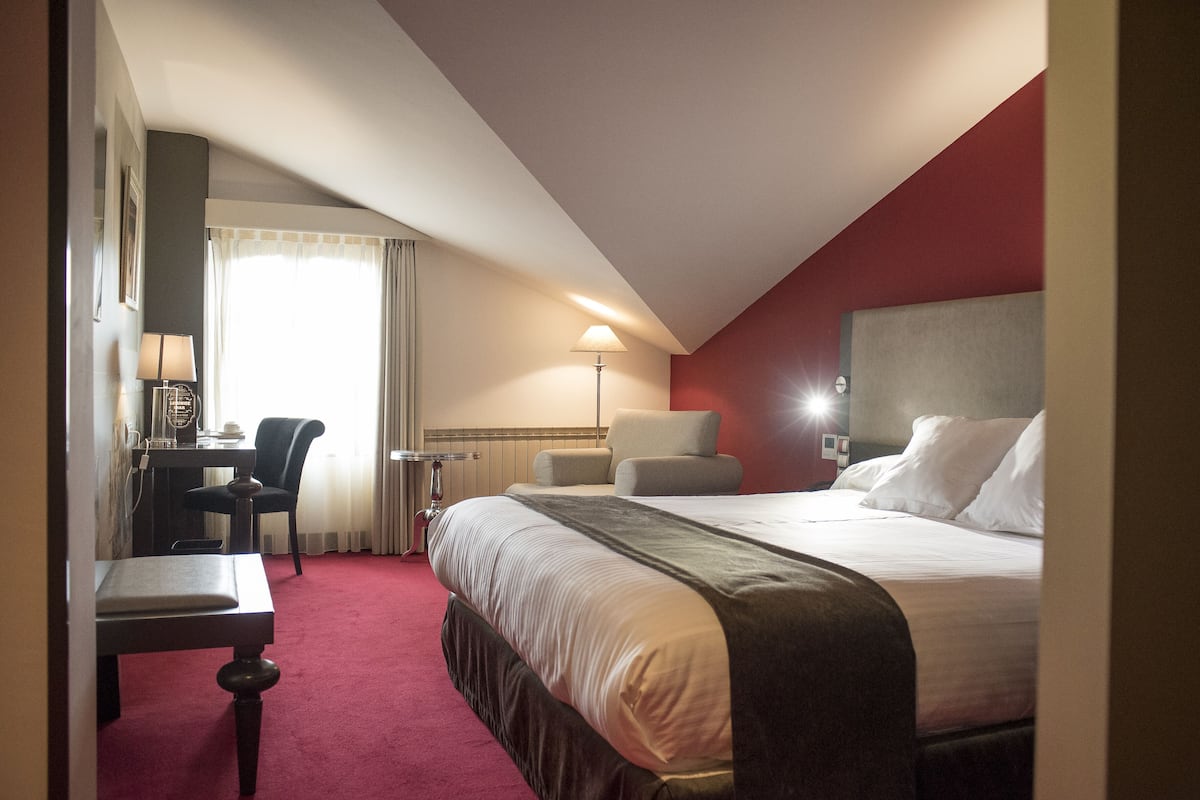A hotel room with a bed, a desk, a chair, a lamp, and a window with curtains.