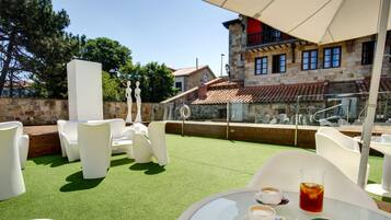 Un área de estar al aire libre con sillas y mesas blancas, una mesa de vidrio con una bebida y pasteles, y un edificio de piedra con techo de tejas rojas al fondo.
