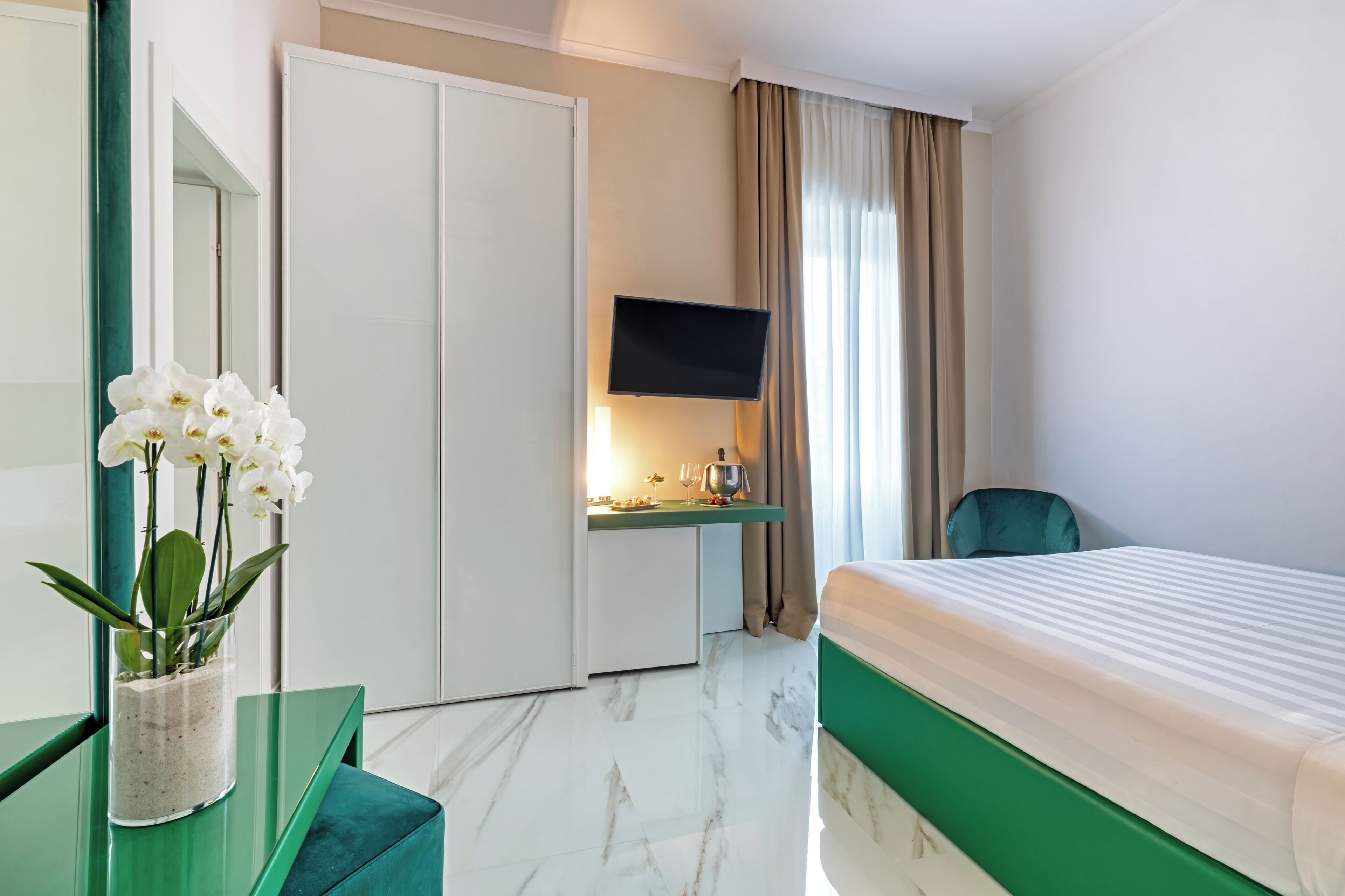 Habitación de hotel con cama, escritorio con televisor de pantalla plana, silla, espejo y un jarrón con flores blancas.