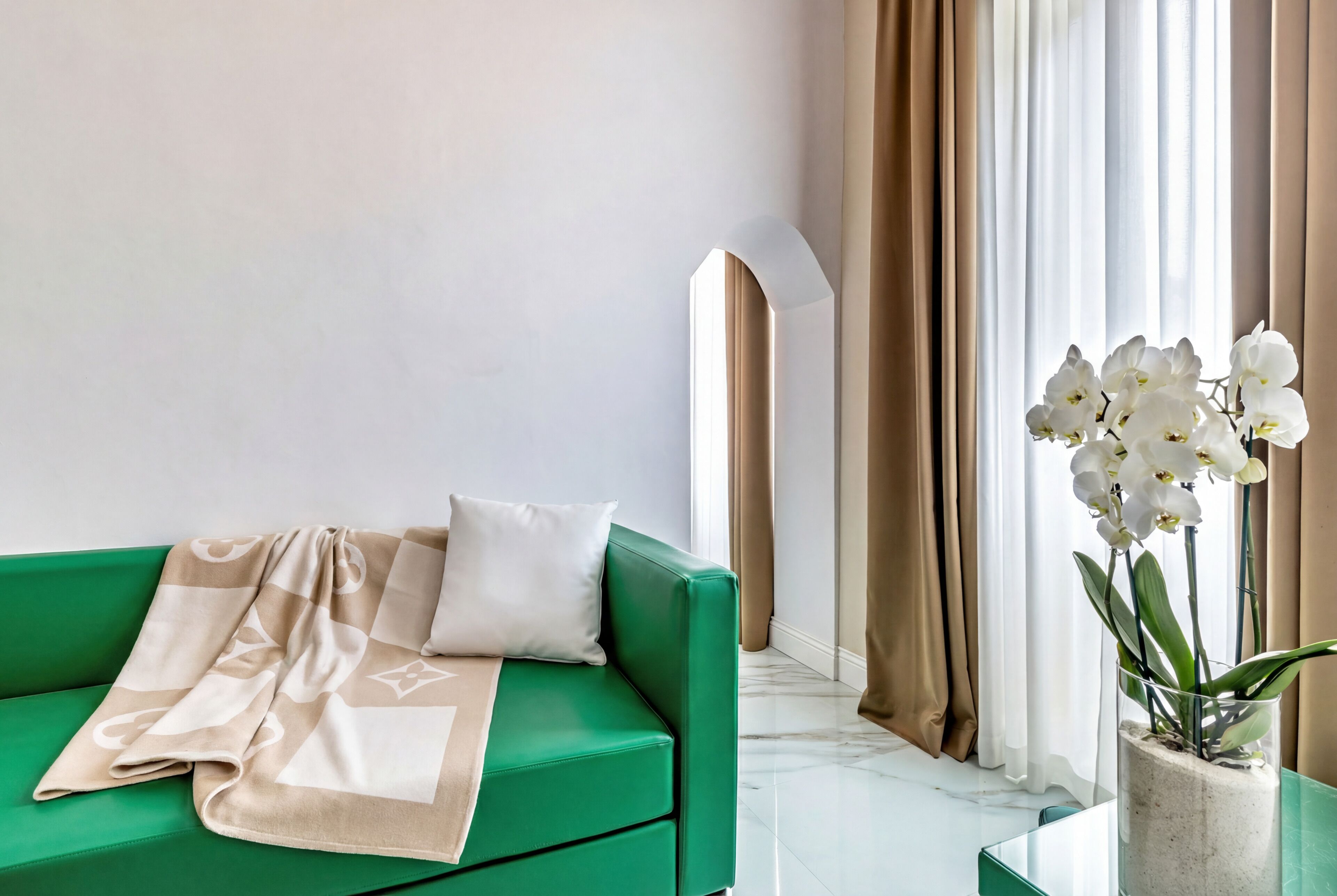 Un sofá verde con una manta beige, una almohada blanca y una jarra de cristal con orquídeas blancas.