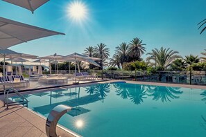 A clear blue pool with sun umbrellas and lounge chairs, surrounded by palm trees.