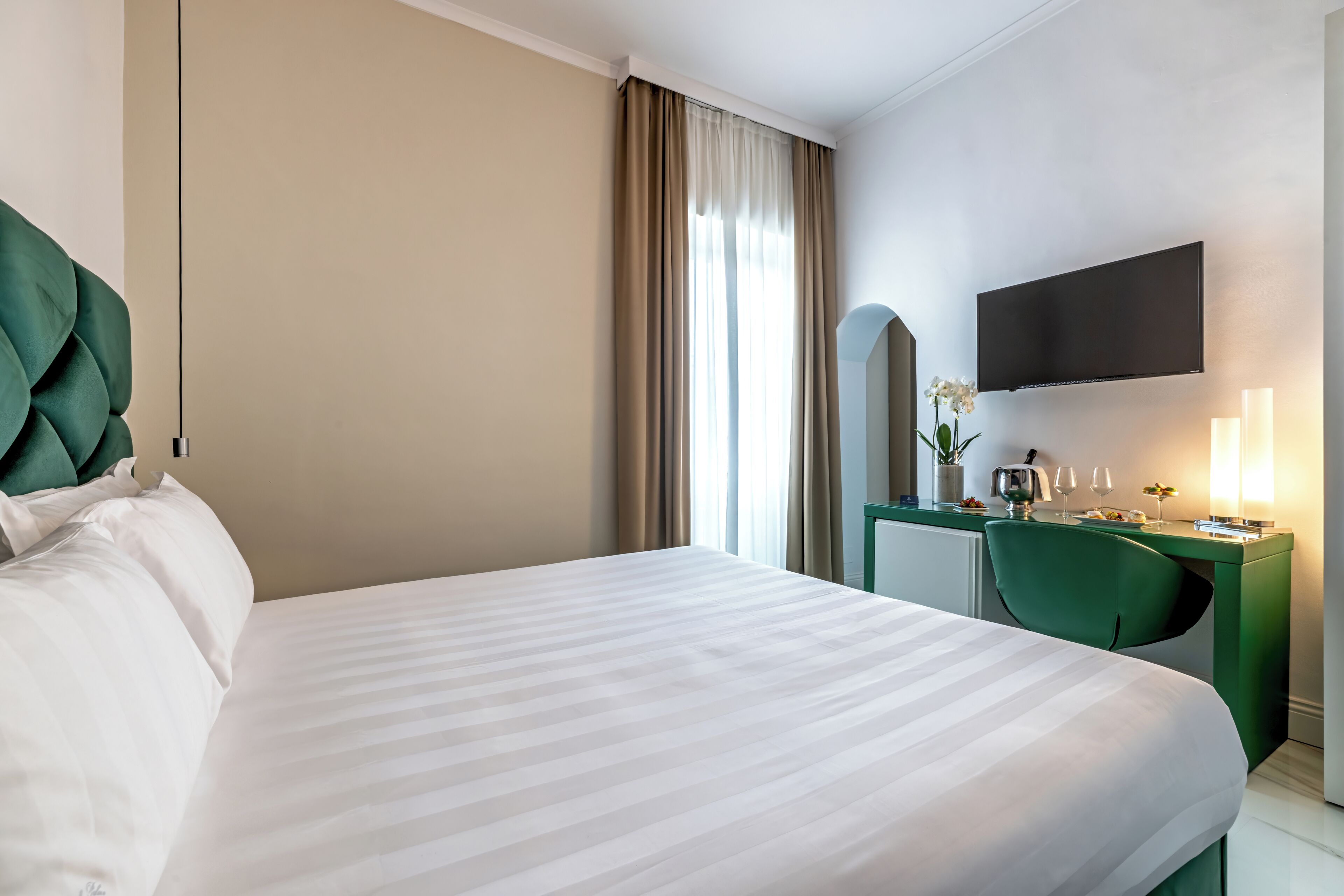 Una camera d'albergo con un letto, una scrivania, una sedia, una televisione e una finestra con tende.